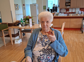 Eine ältere Frau sitzt an einem Tisch in einem gemütlichen Raum. Sie hält ein Stück Kuchen in der Hand und schaut in die Kamera. Vor ihr liegt eine Zeitung und eine Tasse Kaffee steht ebenfalls auf dem Tisch.