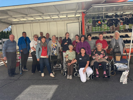 Gruppenfoto von älteren Menschen und Betreuern vor einem Verkaufsstand im Freien. Die Seniorinnen und Senioren sitzen in Rollstühlen oder stehen und lächeln in die Kamera. Im Hintergrund sind Schachteln mit Schuhen sichtbar.