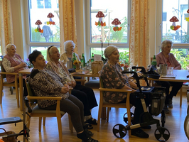 Eine Gruppe älterer Frauen sitzt in einem Gemeinschaftsraum und schaut aufmerksam.
