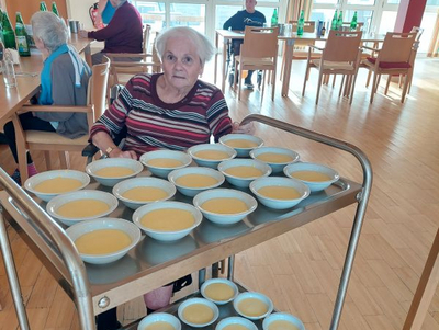 Eine ältere Frau fährt ein Servierwagen mit vielen Schalen Suppe.