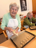 Eine lächelnde ältere Dame mit grauen Haaren in einem hellen Pullover, die einen Spritzbeutel in der Hand hält, während sie Kekse auf ein Backblech mit Backpapier spritzt. Im Hintergrund sitzt eine weitere Frau mit grauen Haaren in einem gestreiften Pullover.