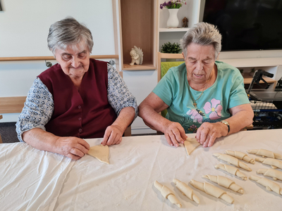 Zwei ältere Frauen sitzen an einem Tisch und rollen Teig. Sie konzentrieren sich auf ihre Arbeit, während vor ihnen bereits einige fertig geformte Teigstücke liegen. Die Szene vermittelt eine Atmosphäre von Gemeinschaft und handwerklicher Tätigkeit.