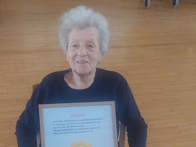 Eine ältere Frau hält eine Urkunde mit einem Goldenen Medaillon in der Hand.
