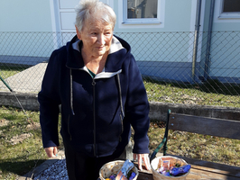 Eine ältere Frau mit grauen Haaren steht im Freien neben einer Bank. Sie trägt einen dunkelblauen Kapuzenpullover und hat zwei kleine Korb mit bunten Gegenständen auf der Bank. Hinter ihr ist ein Gebäude mit Fenstern und eine gepflasterte Fläche zu sehen.