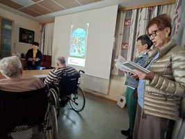 Eine Gruppe von älteren Menschen in einem Raum. Eine Frau steht an der Wand und liest aus einem Buch, während eine Präsentation im Hintergrund gezeigt wird. Einige Personen sitzen im Vordergrund, darunter auch Menschen in Rollstühlen.