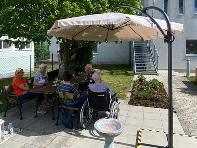 Ältere Menschen sitzen unter einem Sonnenschirm im Garten und genießen die Sonne.