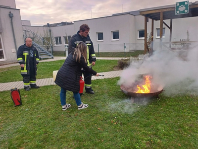 Eine Frau löscht ein Feuer mit einem Feuerlöscher, Feuerwehrleute begleiten sie.
