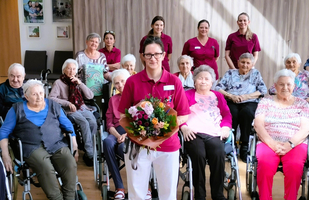 Gruppenszene mit Pflegepersonal und Senioren im Aufenthaltsraum, Blumenstrauß in der Mitte.