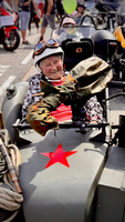 Ein älterer Mann sitzt in einem Motorradgespann und lächelt. Er trägt eine Lederhaube und eine Jacke mit Schulterpolstern. Im Hintergrund sind weitere Motorräder und Menschen zu sehen. Ein roter Stern ist auf dem Gespann sichtbar.