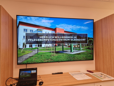 Ein Bildschirm zeigt eine Außenansicht des Pflegekompetenzzentrums Olbendorf mit dem Willkommensslogan. Im Vordergrund stehen einige Bäume und ein Weg, dahinter ist das Gebäude mit einer modernen Architektur zu sehen.