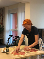 Eine Person mit roten Haaren bereitet Pizza vor. Sie verteilt Belag auf einer Pizza, während weitere Pizzen auf dem Tisch liegen. Im Hintergrund steht eine Flasche Öl und ein Rollstuhl ist sichtbar.
