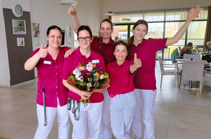 Fünf Mitarbeiterinnen in pinken Oberteilen zeigen einen Blumenstrauß und Daumen hoch.