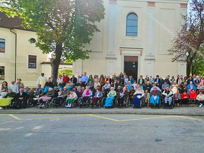 Eine große Gruppe von Menschen, darunter viele im Rollstuhl, steht vor einem Gebäude mit einer weißen Fassade und einer Kirche im Hintergrund. Die Personen sind vielfältig gekleidet und scheinen an einer Veranstaltung oder Versammlung teilzunehmen. Es ist ein sonniger Tag.