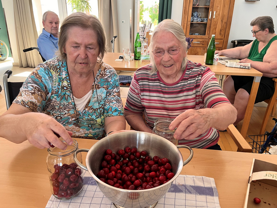 Zwei ältere Frauen sitzen an einem Tisch und füllen Gläser mit Kirschen. Vor ihnen steht eine große Schüssel mit frischen Kirschen. Im Hintergrund sind weitere Personen in einem gemütlichen Raum zu sehen.