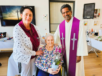 Drei Personen stehen zusammen, eine ältere Frau hält einen Blumentopf mit Orchideen.