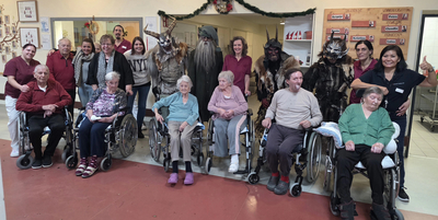 Gruppe von Senioren und Betreuern in Rollstühlen, mit verkleideten Figuren im Hintergrund.