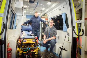 Zwei Männer stehen in einem Krankenwagen, einer sitzt auf der Trage.