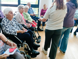 Eine Gruppe älterer Menschen sitzt in einem Raum, einige in Rollstühlen. Eine Person steht und interagiert mit den anderen. Es scheint sich um eine soziale Aktivität oder ein Spiel zu handeln. Ein helles, einladendes Umfeld ist sichtbar.