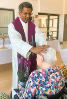 Ein Priester spendet einer älteren Frau den Segen in einem Gemeinschaftsraum.