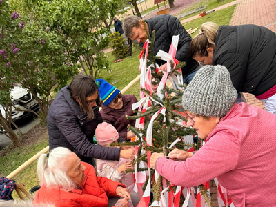 Eine Gruppe von Menschen, darunter Kinder und Erwachsene, dekoriert einen Baum mit roten und weißen Bändern. Einige der Teilnehmer arbeiten aktiv, während andere zuschauen. Die Szene spielt sich im Freien ab, umgeben von grünen Pflanzen und einem gepflasterten Weg.