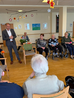 Ein Mann in formeller Kleidung spricht vor einer Gruppe älterer Menschen, die in einem Raum mit sitzenden Stühlen und Dekorationen sitzen. Einige der Anwesenden sind im Rollstuhl. Eine Blumenstrauß befindet sich auf einem Tisch in der Mitte.