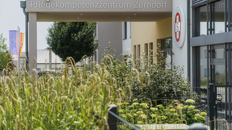 Eingangsbereich des Pflegekompetenzzentrums Zurndorf mit Begrünung im Vordergrund. Über dem Eingang befindet sich ein Schild mit dem Namen der Einrichtung. Im Hintergrund sind Bäume und der moderne Gebäudefront sichtbar.