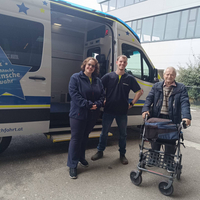 Zwei Mitarbeiter stehen neben einem Transportfahrzeug mit einem älteren Herrn und einem Rollator.