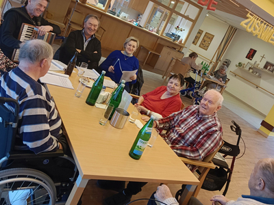 Eine Gruppe älterer Menschen sitzt an einem Tisch in einem Gemeinschaftsraum. Sie lächeln und unterhalten sich, während einige Wasserflaschen auf dem Tisch stehen. Im Hintergrund ist eine festliche Dekoration zu sehen.