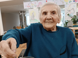 Eine ältere Frau mit einem freundlichen Lächeln sitzt an einem Tisch und hält einen Löffel in einem Glas mit grünem Inhalt. Sie trägt einen blauen Pullover und ist in einem hellen, gut beleuchteten Raum, möglicherweise in einem Gemeinschaftsraum oder Kunststudio. Hintergrund mit bunten Zeichnungen.