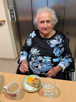 Eine ältere Frau sitzt an einem Tisch und hält ein Besteck in der Hand. Vor ihr steht ein Teller mit Kuchen und eine Tasse mit Kaffee. An der Seite steht ein Glas Wasser. Im Hintergrund ist eine Wand mit einem Aufzug.