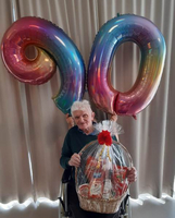 Ein älterer Mann sitzt im Rollstuhl, hält einen Geschenkkorb und feiert 90 Jahre.