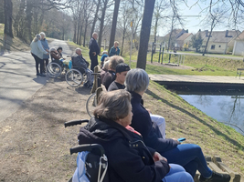 Eine Gruppe von Menschen in Rollstühlen sitzt an einem ruhigen Weg neben einem Gewässer. Im Hintergrund sind mehrere Personen zu sehen, die spazieren gehen. Es ist ein sonniger Tag und die Bäume sind noch ohne Blätter.