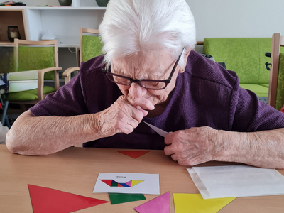 Eine ältere Frau mit Brille sitzt an einem Tisch und betrachtet aufmerksam farbige geometrische Formen. Vor ihr liegen rote, grüne, gelbe, lila und blaue Dreiecke und eine Karte mit einem bunten Muster. Ihr Gesichtsausdruck zeigt Konzentration und Interesse.