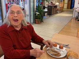 Eine ältere Frau sitzt an einem Tisch mit zwei Eiern und Brot.