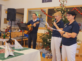 Drei Personen singen und spielen Gitarre vor einem festlich dekorierten Weihnachtsbaum.