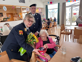Zwei Personen überreichen einer älteren Frau Geschenke und Blumen in einem Gemeinschaftsraum.