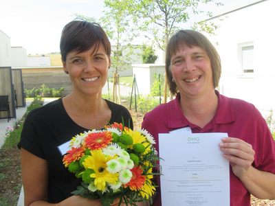 Zwei Frauen halten einen Blumenstrauß und ein Zertifikat in der Hand.