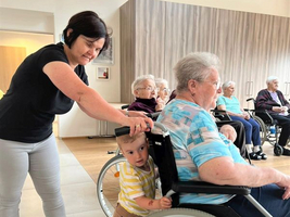 Eine Frau schiebt eine rollstuhlfahrende ältere Dame, während ein Kind neugierig zuschaut.