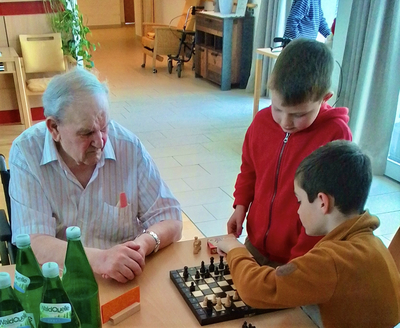 Zwei Kinder spielen Schach mit einem älteren Mann in einem freundlichen Raum.
