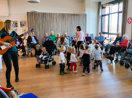 Eine Musikerin spielt Gitarre für tanzende Kinder und Senioren in einem Raum.