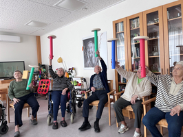 Ältere Menschen heben bunte Stäbe in einer Sitztätigkeit an.