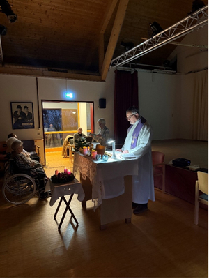 Ein Priester leitet eine Feier mit Kerzenlicht und Teilnehmern in einem Raum.