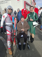 Drei Personen, darunter ein älterer Mann im Rollstuhl, posieren zusammen festlich.