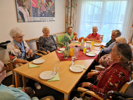 Eine Gruppe älterer Menschen sitzt um einen Tisch, auf dem Teller und Getränke stehen. Sie unterhalten sich freundlich in einer hellen, einladenden Umgebung. Im Hintergrund ist ein großes Kunstwerk zu sehen.