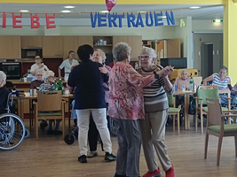 Eine fröhliche Atmosphäre in einem Gemeinschaftsraum, in dem mehrere ältere Menschen tanzen. Zwei Frauen in der Mitte tanzen eng umschlungen, während andere Bewohner auf Stühlen sitzen oder beobachten. An der Wand hängen bunte Banner mit den Wörtern 