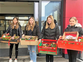 Vier Mädchen halten Pflanzenarrangements in Kartons und Kästen vor einer Tür.