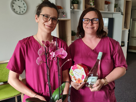 Zwei Frauen in lila Arbeitskleidung stehen in einem Raum. Die eine hält einen Topf mit einer Orchidee, die andere eine Flasche und ein Geschenk. Beide lächeln und stehen in freundlicher Atmosphäre. Im Hintergrund sind Pflanzen und eine Uhr sichtbar.