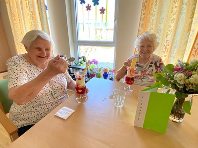 Zwei ältere Frauen genießen Eisbecher in einem hellen Raum.