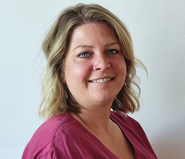 Eine Frau mit blonden, lockigen Haaren steht lächelnd vor einer hellen Wand. Sie trägt ein kurzärmliges, weinrotes Oberteil und hat ihre Hände vor dem Bauch gefaltet.