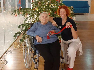 Eine ältere Dame im Rollstuhl posiert mit einer Mitarbeiterin vor einem geschmückten Weihnachtsbaum.